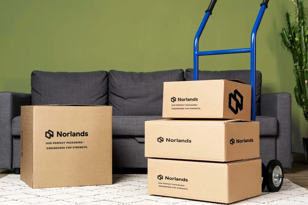 Corrugated Boxes Supplier in Canada.A stack of Norlands-branded cardboard boxes on a hand truck beside a sofa, showcasing durable, eco-friendly packaging for moving or deliveries.