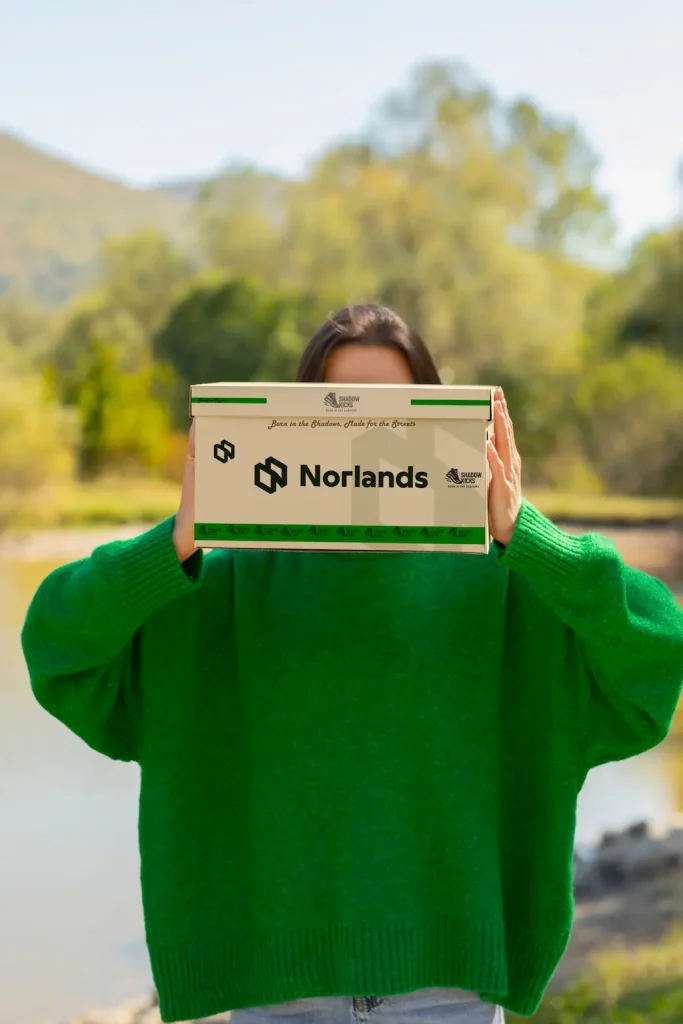 A person in a bright green sweater holds a cardboard box in front of their face. The box is branded with the Norlands logo, slogan “Born in the Shadows, Made for the Greats,” and green decorative accents.