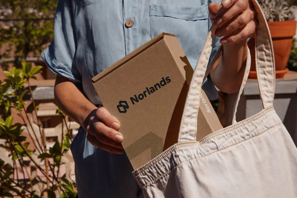 A person places a Norlands-branded cardboard box into a light beige fabric tote bag. The person is wearing a light blue shirt, and the scene is outdoors in natural sunlight.