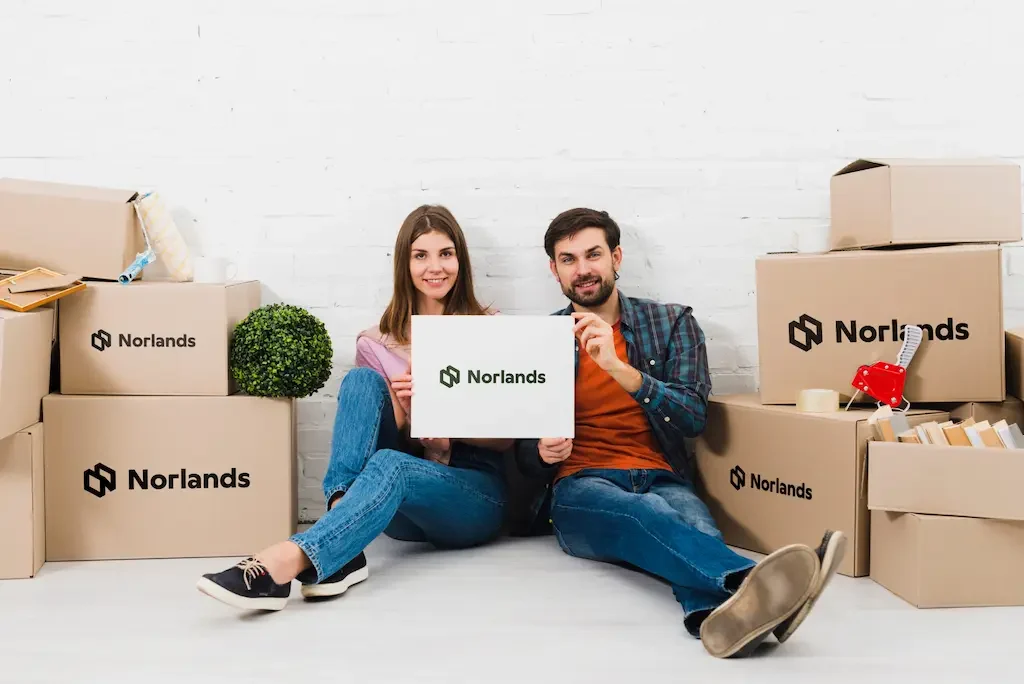 A smiling couple sitting on the floor surrounded by Norlands branded cardboard boxes, holding one box together in front of them.