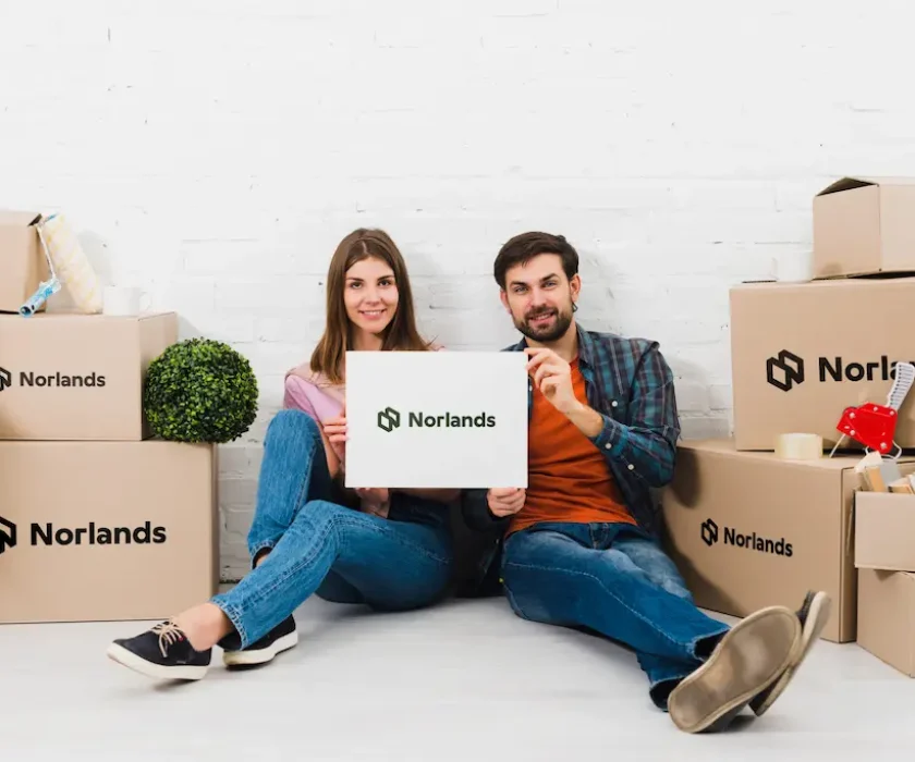 A smiling couple sitting on the floor surrounded by Norlands branded cardboard boxes, holding one box together in front of them.