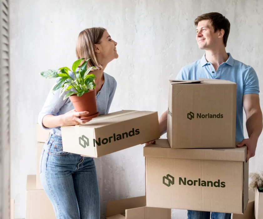 A smiling couple carrying Norlands-branded moving boxes and a potted plant, symbolizing stress-free relocation with durable packaging.