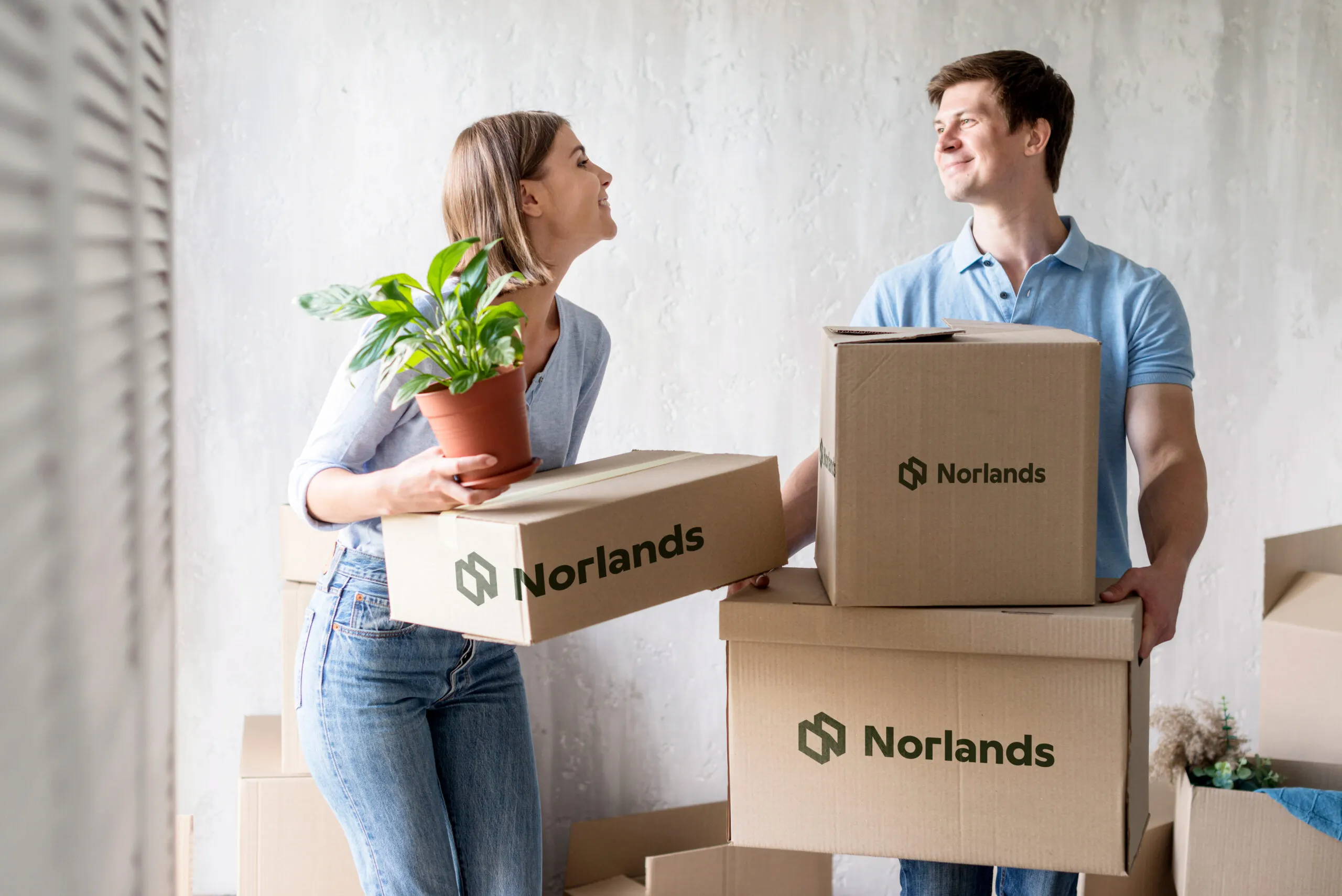 A smiling couple carrying Norlands-branded moving boxes and a potted plant, symbolizing stress-free relocation with durable packaging.