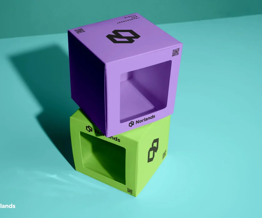 Two modern Norlands packaging boxes in bright purple and green with transparent display windows, stacked on a turquoise surface under studio lighting.