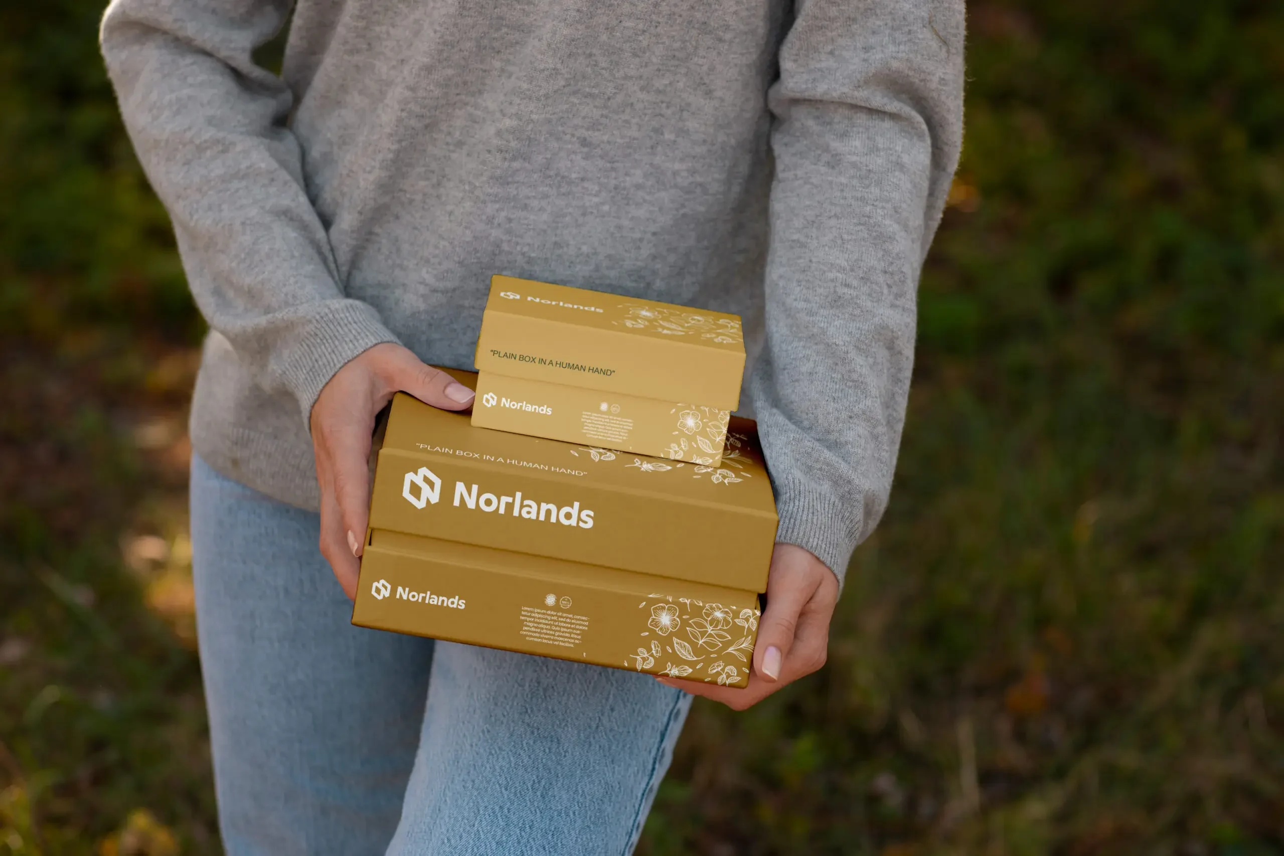 Vancouver custom boxes- Person holding two golden Norlands packaging boxes with floral illustrations, outdoors in natural light.