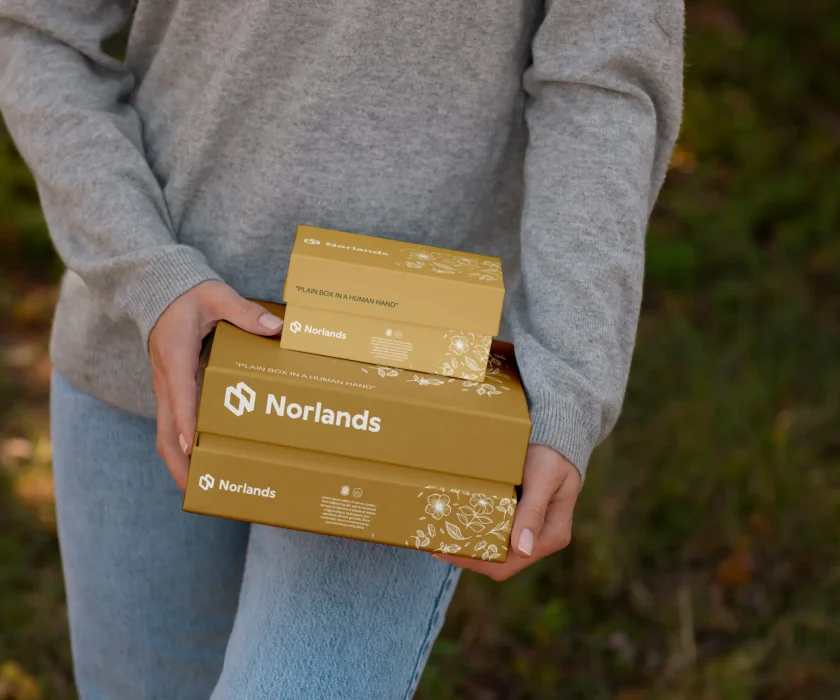 Vancouver custom boxes- Person holding two golden Norlands packaging boxes with floral illustrations, outdoors in natural light.