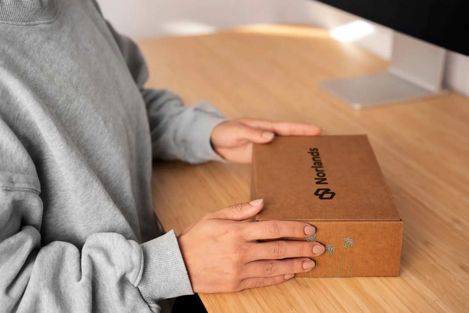 Person in a grey sweater holding a brown Norlands-branded cardboard box on a wooden desk.