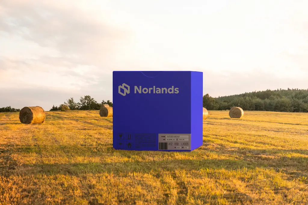 packaging design in Vancouver A vibrant blue NORLANDS box sits in a golden field at sunset, surrounded by hay bales under a warm sky.
