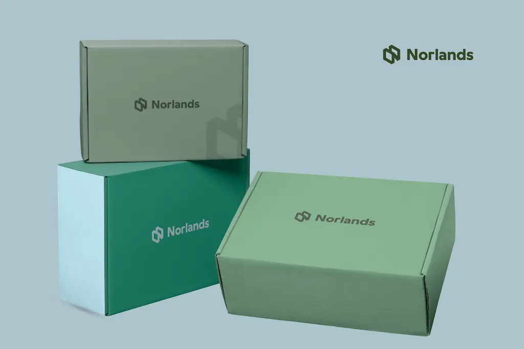 Alt Text:Norlands-branded cardboard box and beige drawstring pouch placed on gravel beside a tree trunk. 